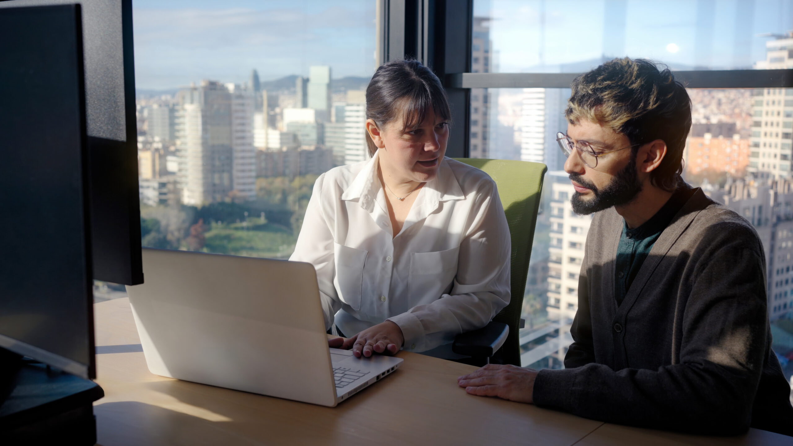Two colleagues discussing work beside a laptop in a sunlit office.